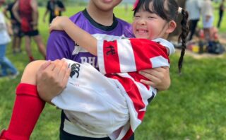 学生ラガールに抱き上げられ笑顔を見せる幼児ラガール。ラグビーファミリーの温かい絆が育まれている。
